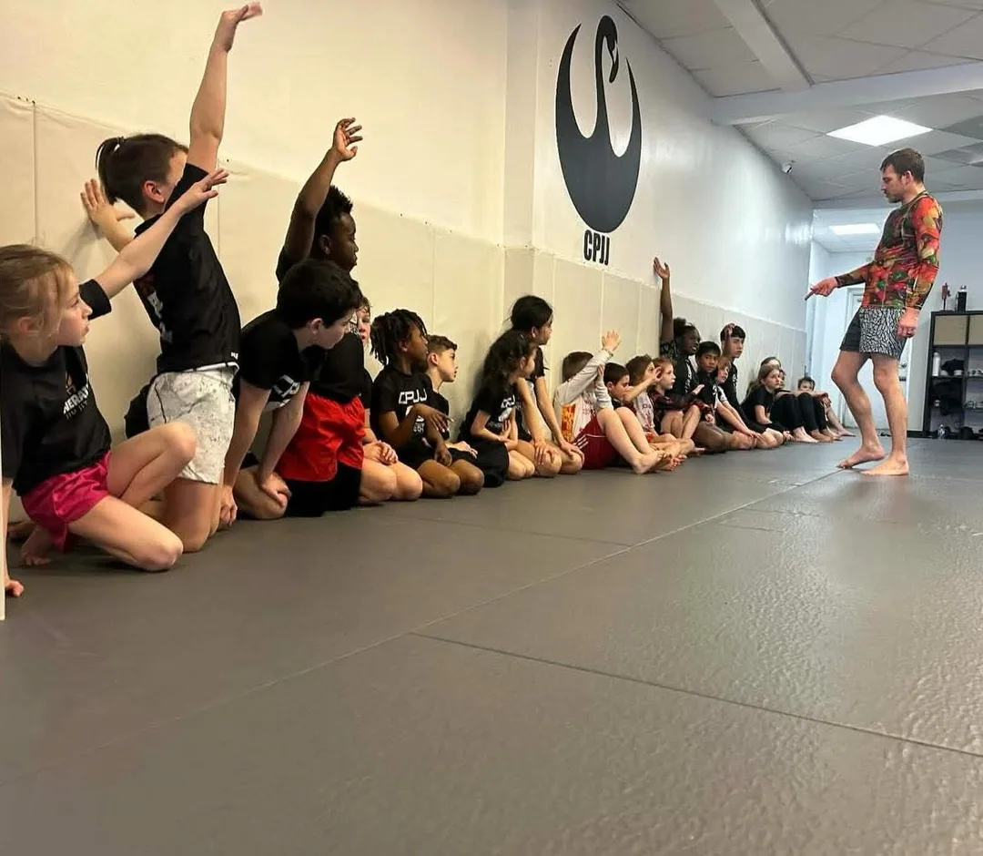Kids kneeling in a line along the mat wall with hands raised to answer a coach during a jiu jitsu class