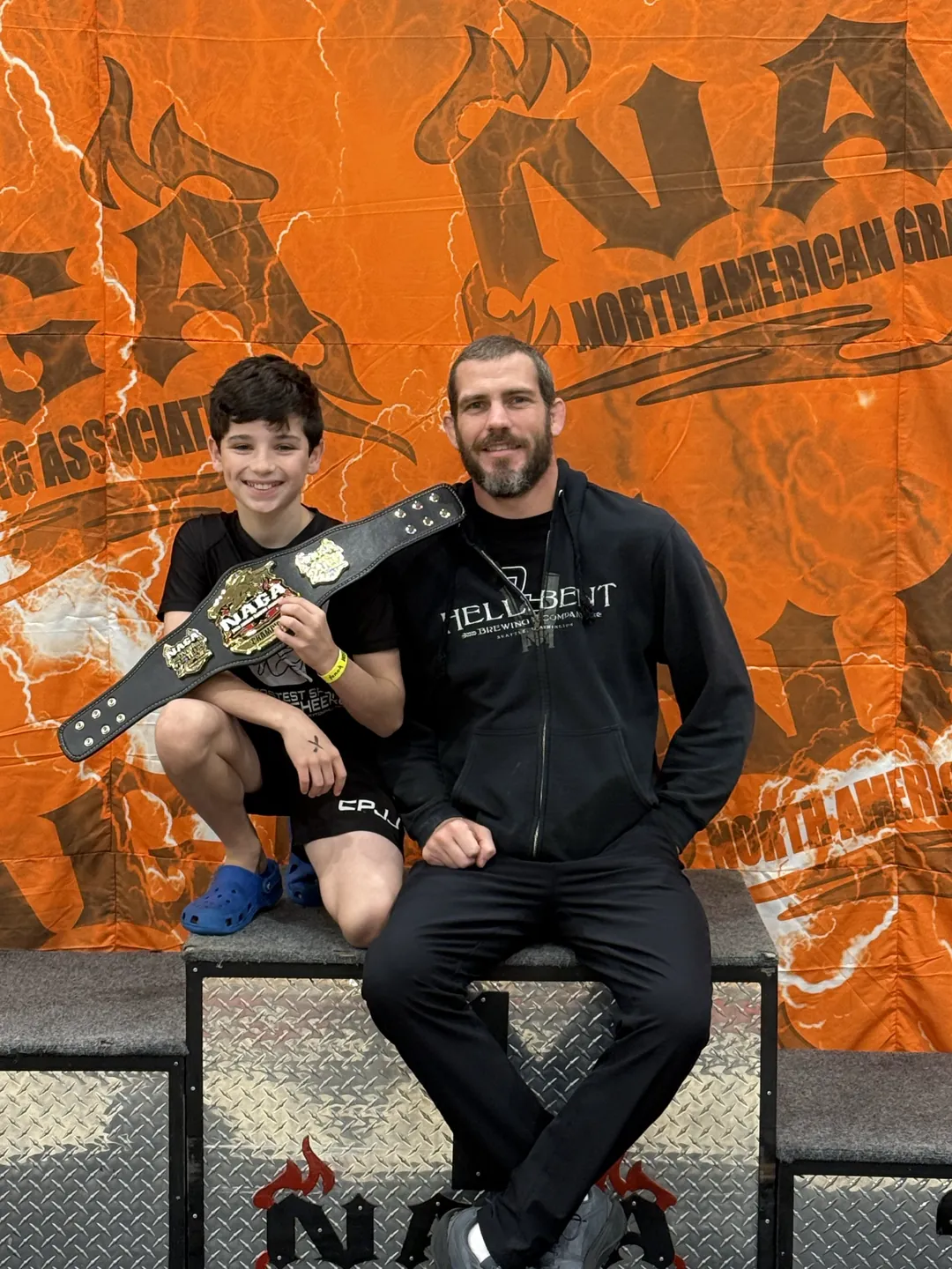 A CPJJ young athlete holding his NAGA championship belt on the podium with coach Clayton Phillips