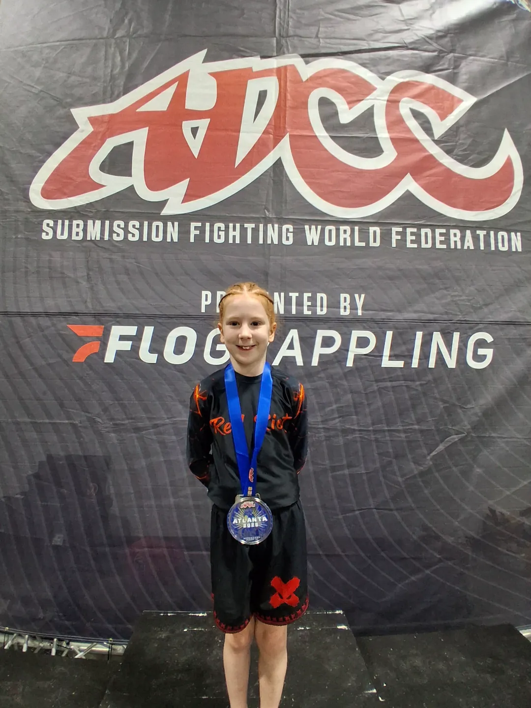 A CPJJ young athlete with her medal in front of the ADCC banner at a submission grappling tournament
